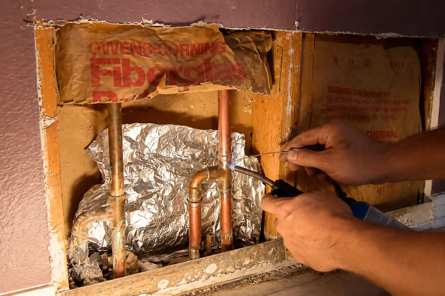 Technician soldering a copper water line inside an opened wall cavity during a slab leak repair
